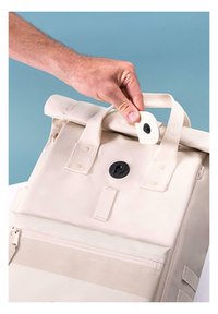Roll-top backpack in beige with a black button closure, canvas material, two top handles, and a front zippered pocket.