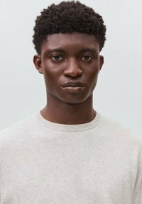 Hellgrauer Pullover mit rundem Ausschnitt und Bündchenmuster, getragen von einer Person mit kurzem, strukturiertem dunklem Haar und neutralem Hintergrund.