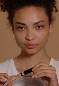 KIKO Concealer mit einem beigen Tubus und schwarzem Deckel, gehalten von einer Person. Der Hintergrund ist ein sanftes Braun. Die Haut ist natürlich mit minimalem Make-up.