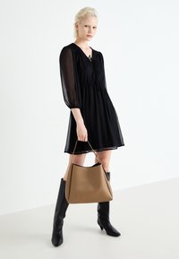 Crna, lepršava haljina s puf rukavima i vezanjem oko vrata, uparena s velikom bež torbom i crnim čizmama do koljena. Glatke teksture kroz cijeli outfit.
