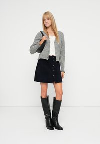 Mulher vestindo um cardigã cinza, blusa branca, saia preta com botões na frente, botas altas pretas até o joelho e carregando uma bolsa de ombro preta, de pé contra uma parede branca.