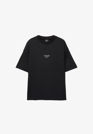 Schwarzes T-Shirt mit kurzen Ärmeln und weißem Text "In der Mitte von Nirgendwo" zentriert auf der Brust, Rundhalsausschnitt und lockere Passform.