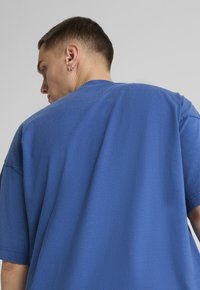 Modré oversized tričko viděné zezadu, s krátkými rukávy, kulatým výstřihem a hladkou, mírně texturovanou tkaninou.