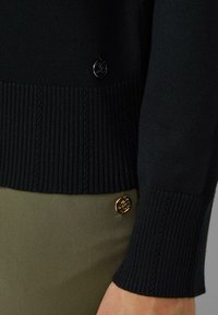 Zwarte gebreide trui met geribde zoom en manchetten, versierd met decoratieve gouden logo knopen nabij de zoom, gecombineerd met olijfgroene stof.
