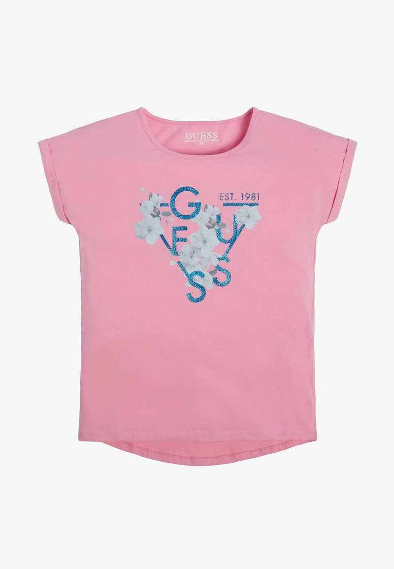 T-shirt rose à manches courtes avec nom de marque pailleté bleu "GUESS,", imprimé floral blanc, et texte "EST. 1981" à l'avant.