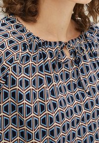 Kvinde iført en bluse med geometrisk mønster i nuancer af blå, sort og brun, med en rund halsudskæring og en knyttet sløjfedetalje.