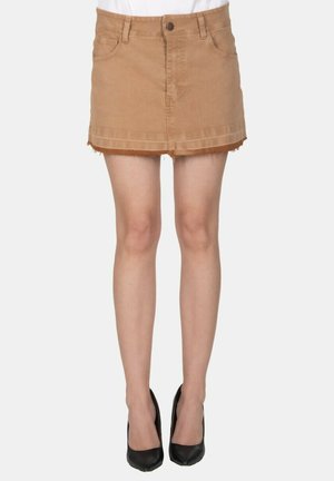 Minigonna di denim color cuoio con orlo grezzo, indossata con décolleté nere a punta su gambe pallide su sfondo bianco.