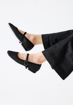 Zapatos planos de malla negra con punta afilada, que presentan una pequeña correa con hebilla y un tacón bajo y cuadrado. Combinados con pantalones oscuros de pierna ancha.