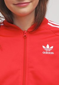 Červená zipová bunda vyrobená z hladké tkaniny, s bílým logem adidas a třemi bílými pruhy na ramenou. Detail zipu v přední části.