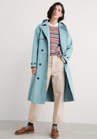 Jasnoniebieski płaszcz trench z podwójnym zapięciem, czarnymi guzikami, w zestawieniu z kolorowym swetrem z dzianiny i beżowymi spodniami. Brązowe buty.