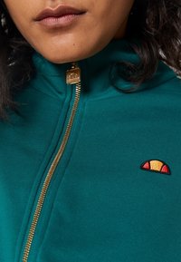 Teal zip-up jacket with gold zipper, featuring a small multi-colored embroidered logo on the left chest area. Soft fabric with a smooth texture.