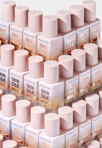 Stapelade flaskor med "Revolution Skin Silk" foundation; fyrkantiga vita baser, ljusrosa lock och guldtextetiketter som indikerar nyanser.