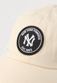 Beżowa czapka z daszkiem z czarno-białą okrągłą naszywką New York Yankees, zawierającą logo "NY" oraz napis "Est. 1903".
