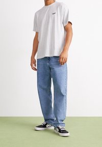 Homme portant un t-shirt gris clair, un jean bleu ample et des baskets à carreaux noir et blanc, se tenant sur un sol vert clair.