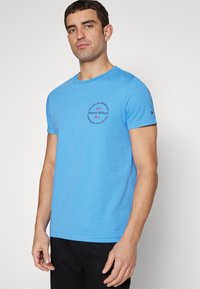 Jasnoniebieski bawełniany t-shirt z krótkim rękawem, okrągłym dekoltem oraz nadrukiem z napisem "Tommy Hilfiger" i "1985" w kolorze czerwonym.