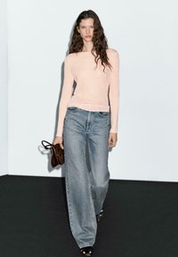 Frau mit langen, gewellten Haaren, die ein blassrosa Langarmoberteil, weite blaue Jeans, schwarze Sandalen trägt und eine kleine braune Handtasche hält.