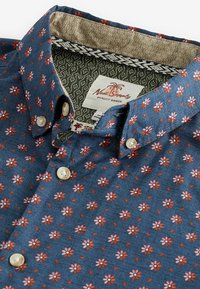 Blauw overhemd met knopen en een klein roze en rode bloemenpatroon, met beige knopen, waardoor de gedetailleerde patroonnvoering van de kraag en het merkembleem zichtbaar zijn.