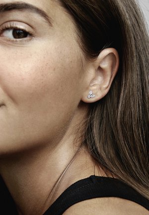 Strieborné náušnice s jedinečným tvarom, zdobené dvoma iskriacimi čírymi drahokamami. Detailný záber zachytáva náušnicu na uchu vedľa hladkej pleti.