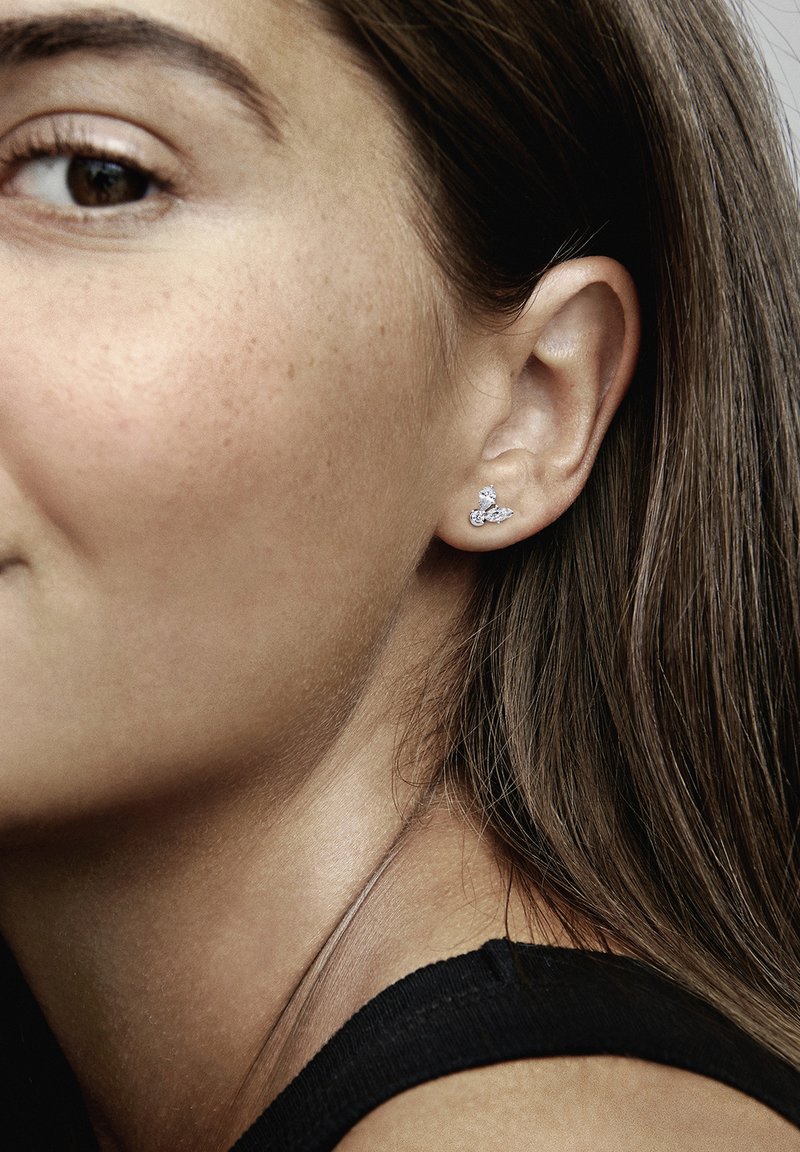 Strieborné náušnice s jedinečným tvarom, zdobené dvoma iskriacimi čírymi drahokamami. Detailný záber zachytáva náušnicu na uchu vedľa hladkej pleti.