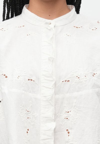 Fehér vászon ing álló gallérral, finom virágos hímzéssel és fodrokkal a gombolósáv mentén. Finom kivágások díszítik.