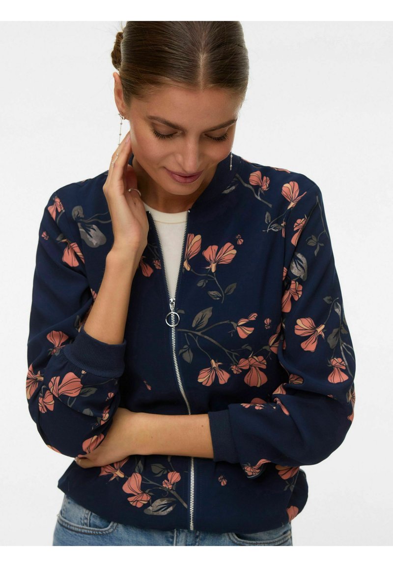 Femme aux cheveux tressés portant une veste zippée bleu marine à fleurs, regardant vers le bas, une main touchant son cou et l'autre croisée sur sa taille.