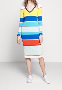 Abito lavorato a maglia a righe con scollo a V, maniche lunghe e silhouette aderente. I colori includono giallo, blu, rosso e azzurro.