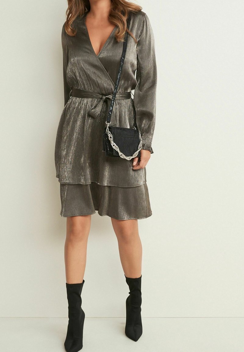 Robe portefeuille bronze scintillante à manches longues avec ceinture à la taille, ourlet à volants en couches, associée à un sac à main noir texturé et des bottines noires.