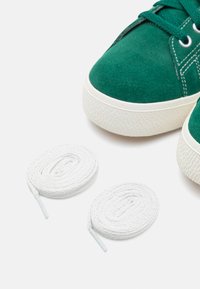 Sneakers verdes em camurça com solas de borracha brancas e detalhes texturizados, acompanhados por dois atacadores brancos planos dispostos em espirais.