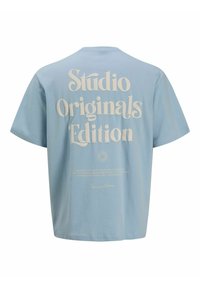 T-shirt bleu clair à manches courtes avec grand texte crème « Studio Originals Edition » dans le dos et texte plus petit avec logo en dessous.