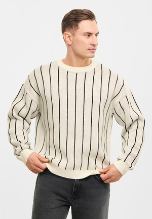 Suéter beige de punto con rayas negras verticales, cuello redondo y puños acanalados, combinado con jeans oscuros. Diseño texturizado y de corte suelto.