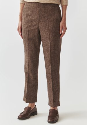 Brązowe, dopasowane spodnie z wełny z mankietami, noszone z brązowymi skórzanymi mokasynami i beżowym swetrem na stojącej osobie.