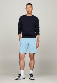 Maglione girocollo navy, pantaloncini azzurri, calze bianche e sneakers nere. Modello in piedi con le mani in tasca, capelli corti.