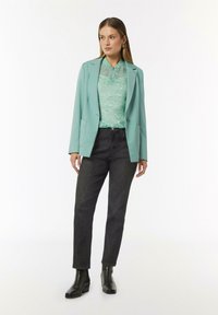 Mintgrüner Blazer über einem gemusterten hellen Oberteil, kombiniert mit dunklen Jeans und schwarzen Knöchelstiefeln. Verfügt über Taschen und einen taillierten Schnitt.