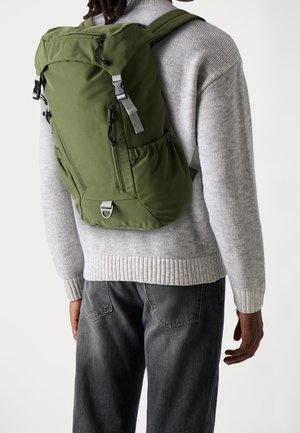 Personne portant un sac à dos vert avec plusieurs fermetures éclair sur un pull gris et un jean noir, vue de dos.