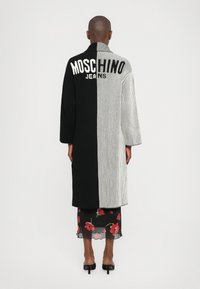 Črno-bela rebrasta bunda z logotipom "MOSCHINO JEANS" na hrbtu. Ima dolg silhuet in široke rokave, nosi se čez cvetlično krilo.