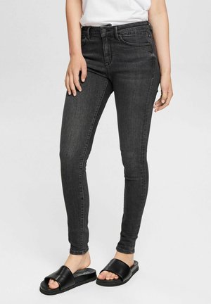 Mujer con vaqueros pitillo gris oscuro, top blanco y sandalias negras de tipo slide, de pie frente a un fondo claro y liso.