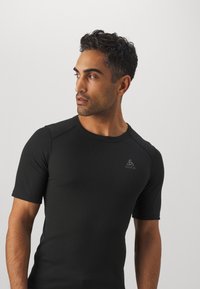 T-shirt de sport noir à manches courtes avec une coupe ajustée, en tissu léger et logo ton sur ton sur le côté supérieur gauche. Texture lisse.