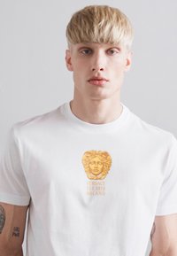 Blond man wearing white Versace t-shirt with gold Medusa logo and text "VERSACE EST. 1978 MILANO" on front, neutral background.