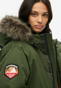 Green parka jacket with a fur-lined hood. Features a patch on the sleeve and a zip closure. Smooth texture, designed for cold weather.