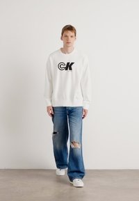 Felpa bianca con logo "CK" nero, abbinata a jeans blu strappati con grandi buchi e sneakers bianche. Look semplice e casual.