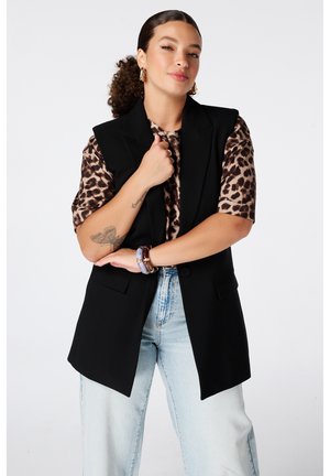 Femme aux cheveux bouclés en queue de cheval portant une chemise imprimé léopard, un blazer noir sans manches, un jean clair, les bras croisés, souriant légèrement.