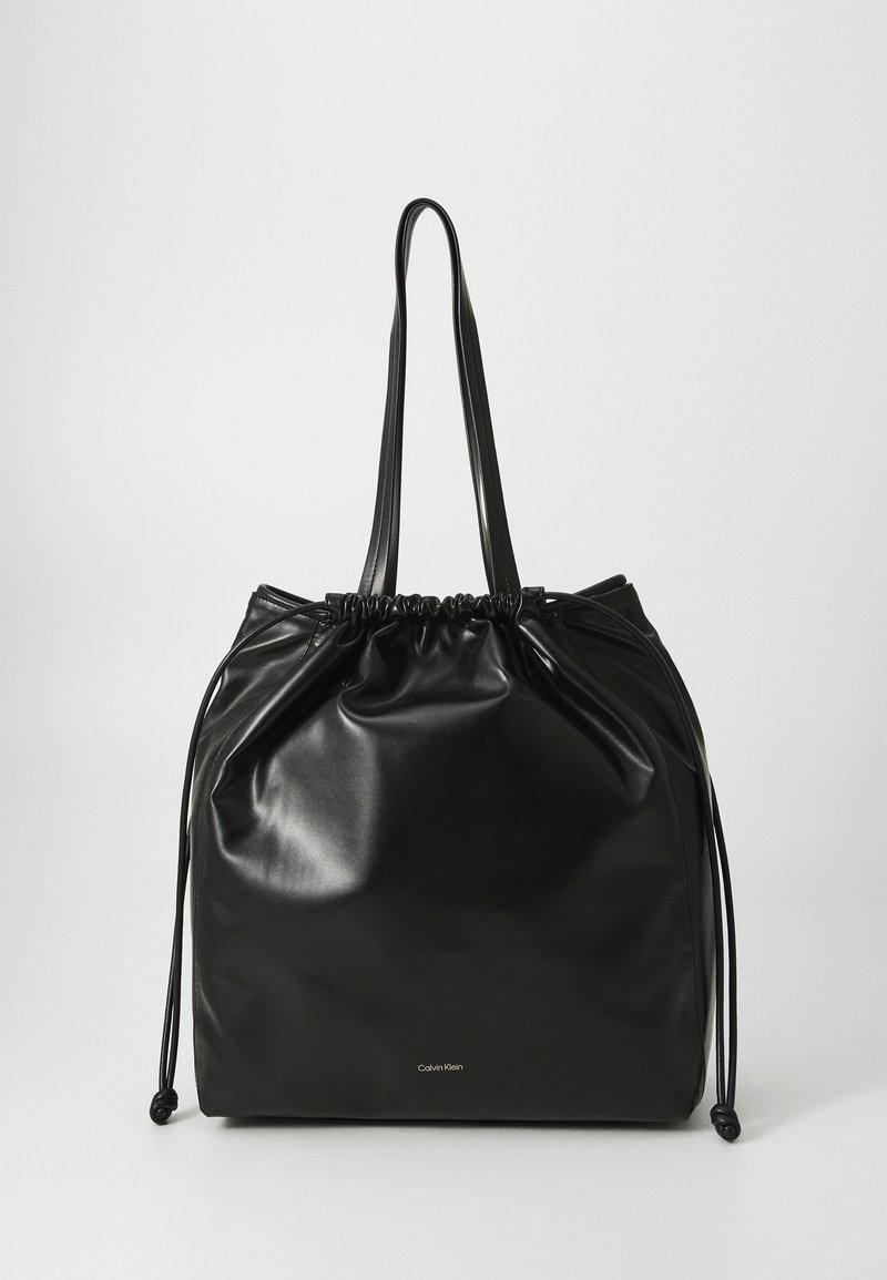 Borsa a tracolla nera con coulisse realizzata in materiale lucido. Presenta manici lunghi e un logo Calvin Klein ben visibile in basso. Design semplice.