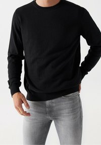 Homme portant un pull noir uni à manches longues et un jean ajusté gris clair, debout devant un fond blanc.