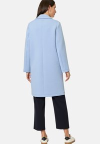 Lichtblauwe, lange, met kraag gemaakte jas met een gladde textuur, zichtbare naden en een subtiel detail aan de achterkant, gecombineerd met donkere cropped pantalons en sneakers.