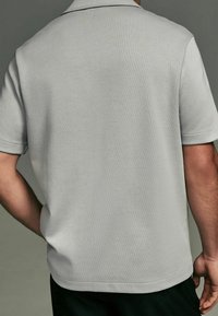 Polo shirt grigio chiaro realizzato in tessuto morbido, con colletto, maniche corte e orlo dritto. Design minimalista senza motivi visibili.