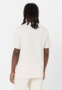 Camiseta de manga corta color crema, hecha de una tela suave, con un corte relajado y pequeños botones de acento en el lado. Se muestra la vista trasera.