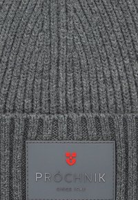 Szary dzianinowy beanie o prążkowanej teksturze. Zawiera skórzaną naszywkę z nazwą marki "PRÓCHNIK" oraz czerwoną emblemat.