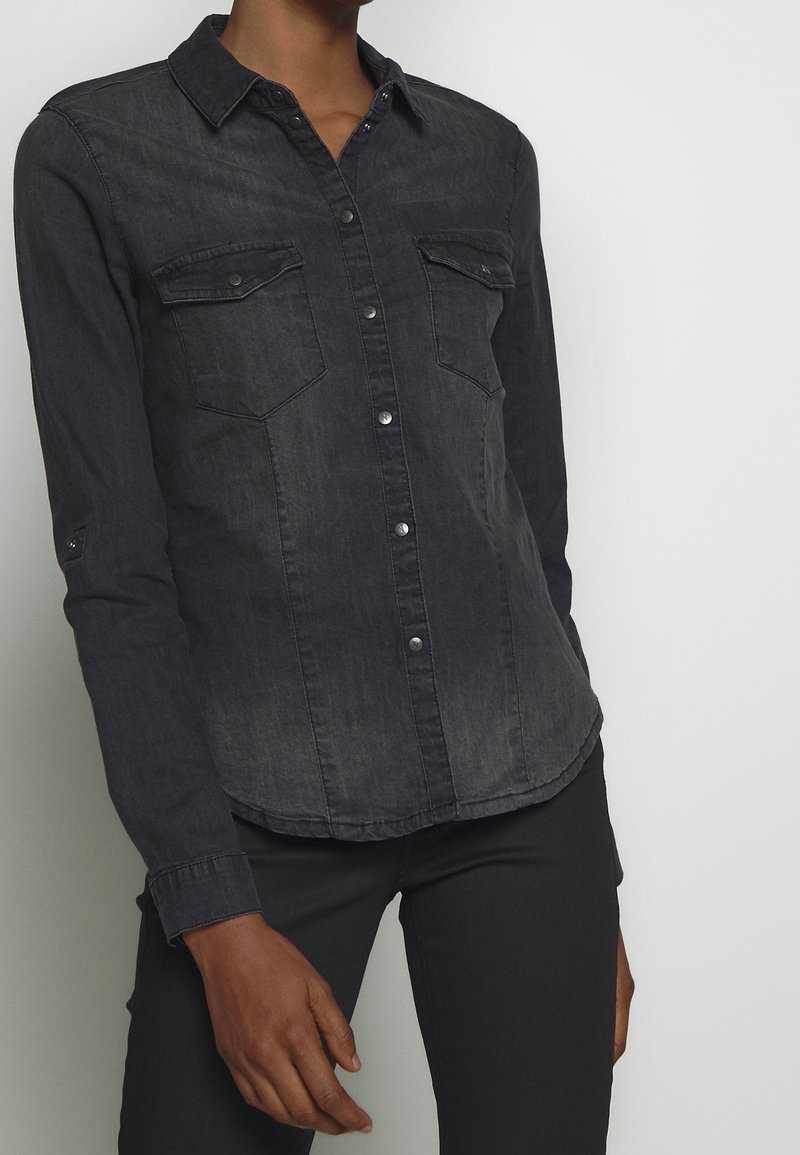 Personne portant une chemise en denim noir ajustée à boutons avec deux poches poitrine et un pantalon noir sur un fond clair uni.