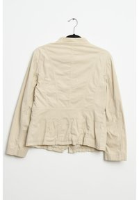 Béžová bavlněná bunda s peplum lemem, vypasovaným pasem a dlouhými rukávy. Má úpravy švů a klasický límec. Visí na černém háčku.