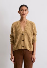 Strickcardigan in hellem Beige mit vertikaler Rippstruktur, tiefem V-Ausschnitt und braunen Knöpfen. Über braunen Wildlederhosen getragen.